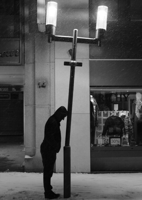 grayscale photo of man in black hoodie standing near glass window